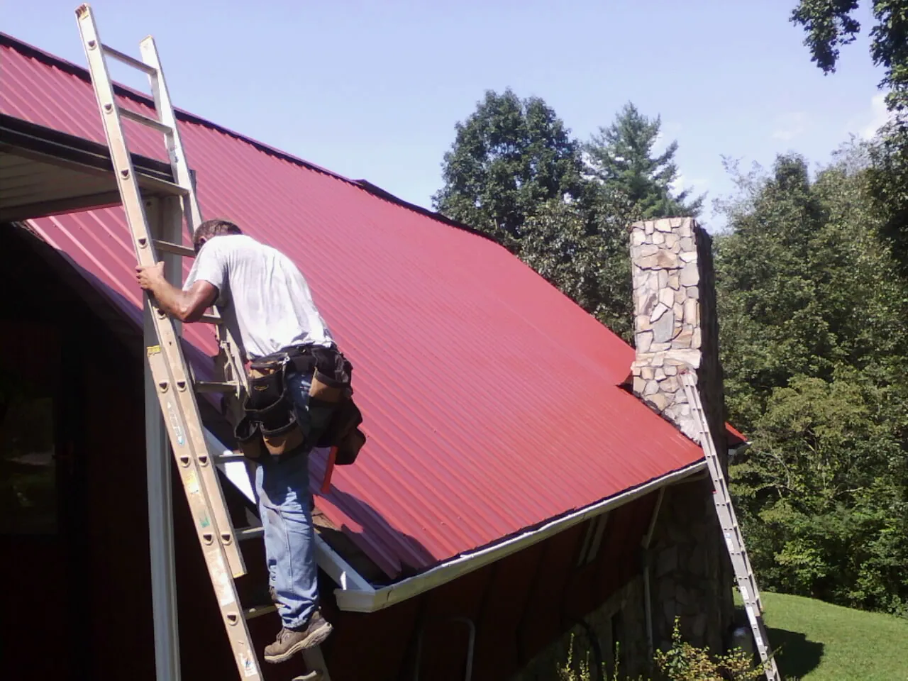 Roofer on a ladder beside a red metal roof and stone chimney — Soffit & Fascia Repair by Roofing Brothers in Houtzdale